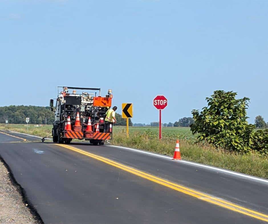 A pavement marking truck with its crew painting lines on a rural highway next to a field.